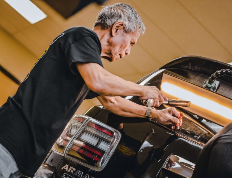 A person engaged in dent repair at the rear of a car, focused on the task at hand with tools in hand.