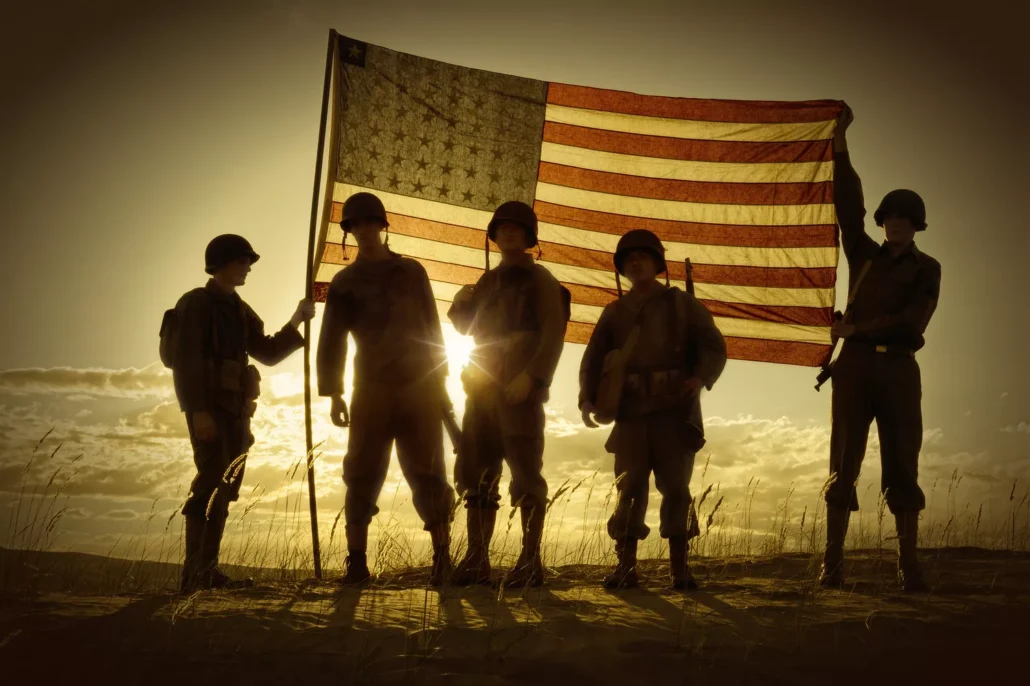 Silhouetted soldiers in vintage uniforms, representing veterans, stand holding a large American flag against a sunset backdrop, creating a dramatic scene.