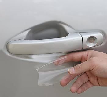 A hand carefully applies a clear PPF underneath the handle of a silver car door, ensuring its protection.