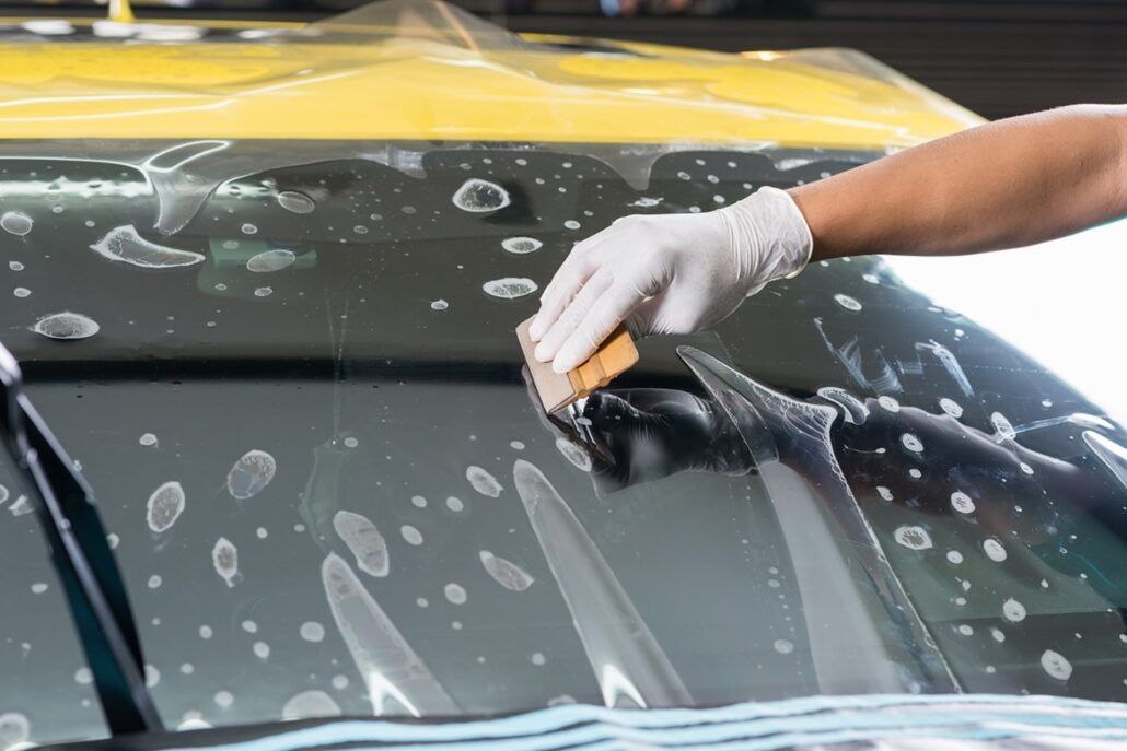 A gloved hand applies a squeegee to a soapy car windshield, ensuring the guard is spotless during cleaning.