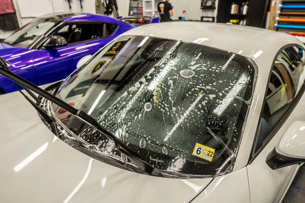 Two cars in a garage, one white and one blue. The white car, equipped with windshield protection, has its wipers lifted, soap suds on the windshield, and the hood raised. The blue car has its hood resting on the windshield.