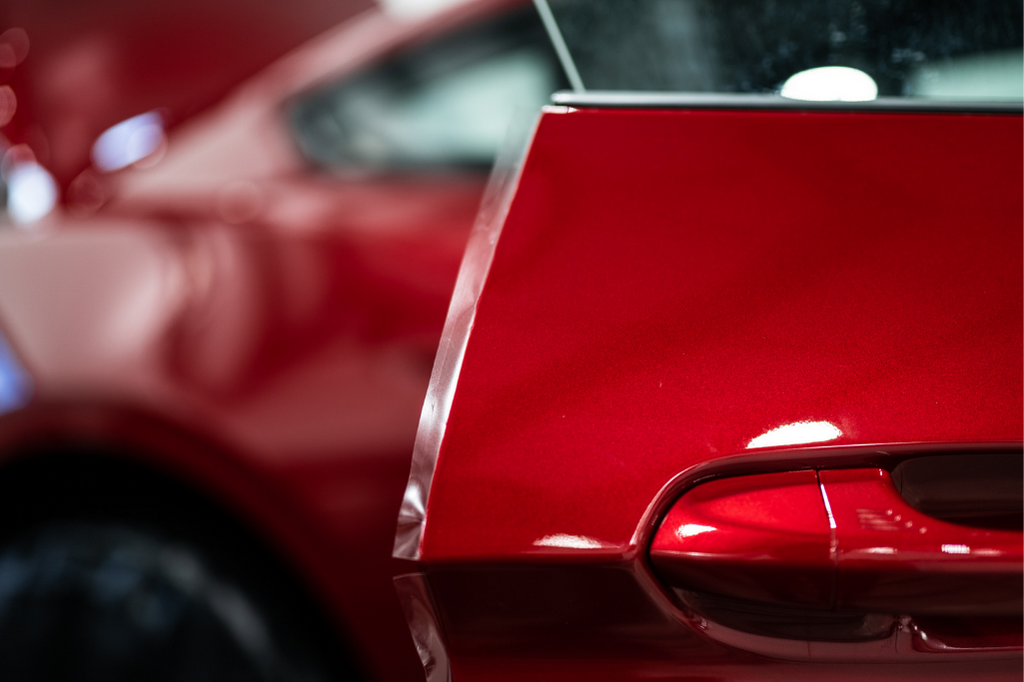 Close-up of a shiny red car door slightly ajar, showing part of the vehicle's rear area and window with a blurry reflection in the background. The sleek design is complemented by an expertly applied PPF, ensuring the pristine condition of every surface.