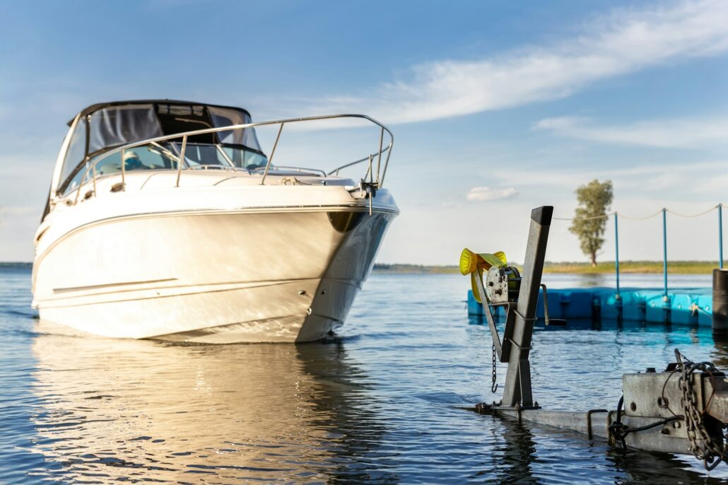 A white motorboat, safeguarded by marine-grade paint protection film, is partially docked in calm water near a small blue dock, with clear skies and some trees visible in the background.