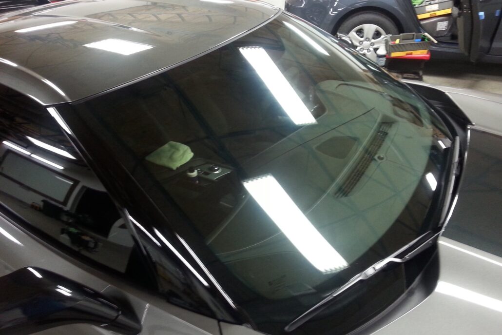 A car's windshield guard and hood shine under bright fluorescent lights, with the reflection visible on the glass. The driver's door is open, showcasing a cleaning cloth on the dashboard.