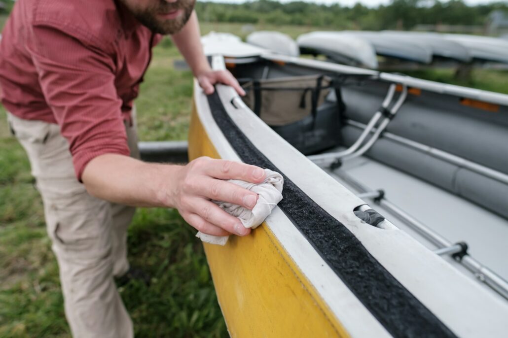 A person in a red shirt and beige pants is cleaning a yellow boat with a cloth. The boat, positioned on grass with other vessels visible in the background, gleams thanks to its marine paint protection.