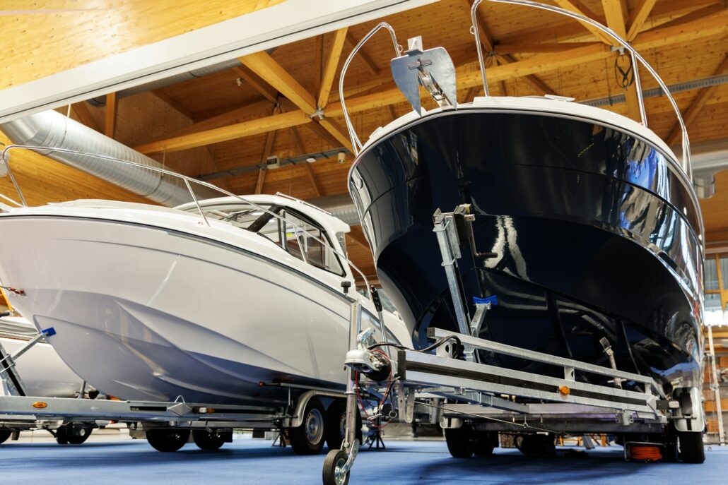 Two boats on trailers are displayed indoors, with wooden ceiling beams visible above. The left boat is white, and the right boat is dark blue. Both Marine vessels, positioned on a blue carpeted floor, boast Paint Protection Film to maintain their pristine condition.