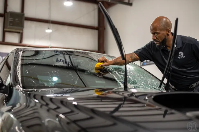 A man cleans a car windshield with a sponge inside a garage, ensuring the PPF overlay stays intact. The car's windshield wipers are lifted.