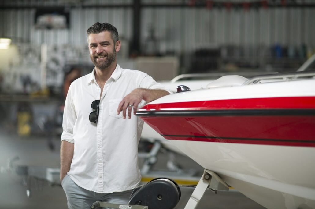 A man with a beard, wearing a light-colored shirt, stands smiling and leaning against a boat inside a workshop. Marine Paint Protection Film is utilized on the boat, shining amidst the busy workspace visible in the background.