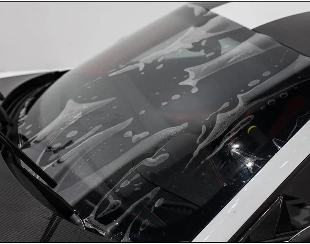 Close-up of a car windshield with soapy water and suds, indicating it is in the process of being washed. The windshield wipers are visible, and the car exterior appears black and white, protected by a PPF film for added durability.