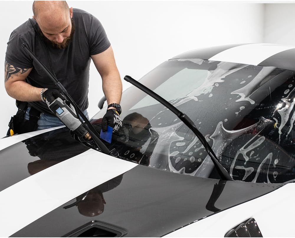 A man installs a protective film on the windshield of a black and white car, smoothing it out with a tool and wearing work gloves. The Windshield Guard ensures durability and clear vision.